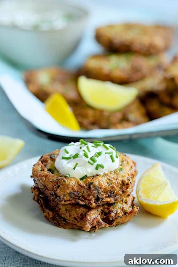 Two Zucchini Ricotta Fritters on a white plate, topped with creamy yogurt and garnished with fresh chives and lemon wedges, ready to be enjoyed.