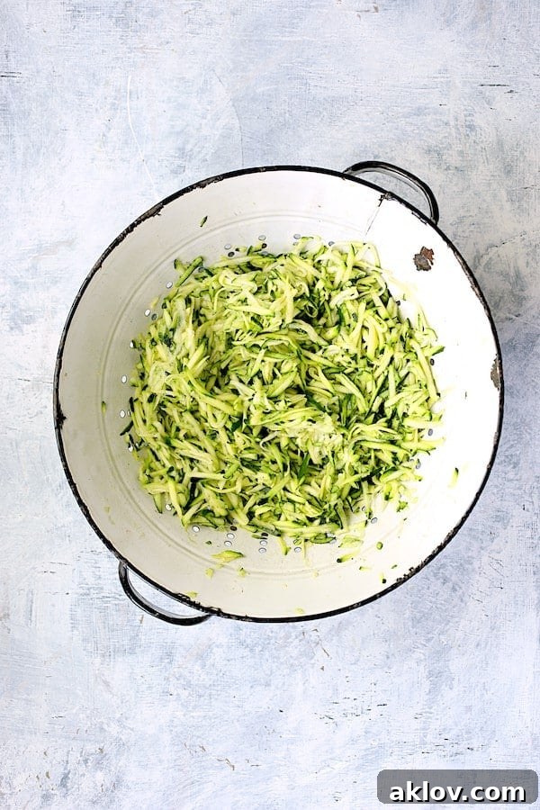 A close-up photo of coarsely shredded zucchini draining in a white colander, showing the moisture being released.