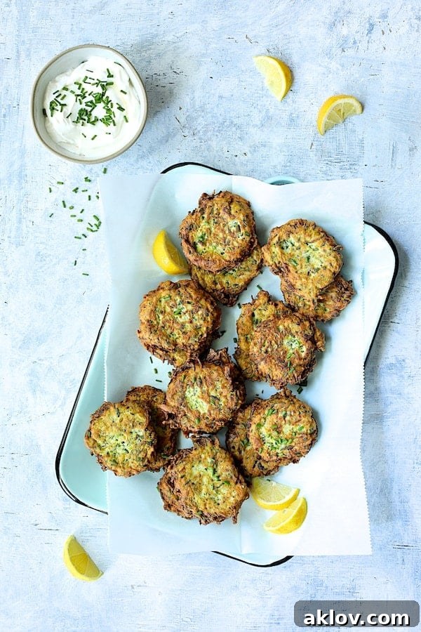 A vibrant serving of Zucchini Ricotta Fritters on a light blue tray, accompanied by a small bowl of creamy yogurt garnished with chives, and fresh lemon slices.
