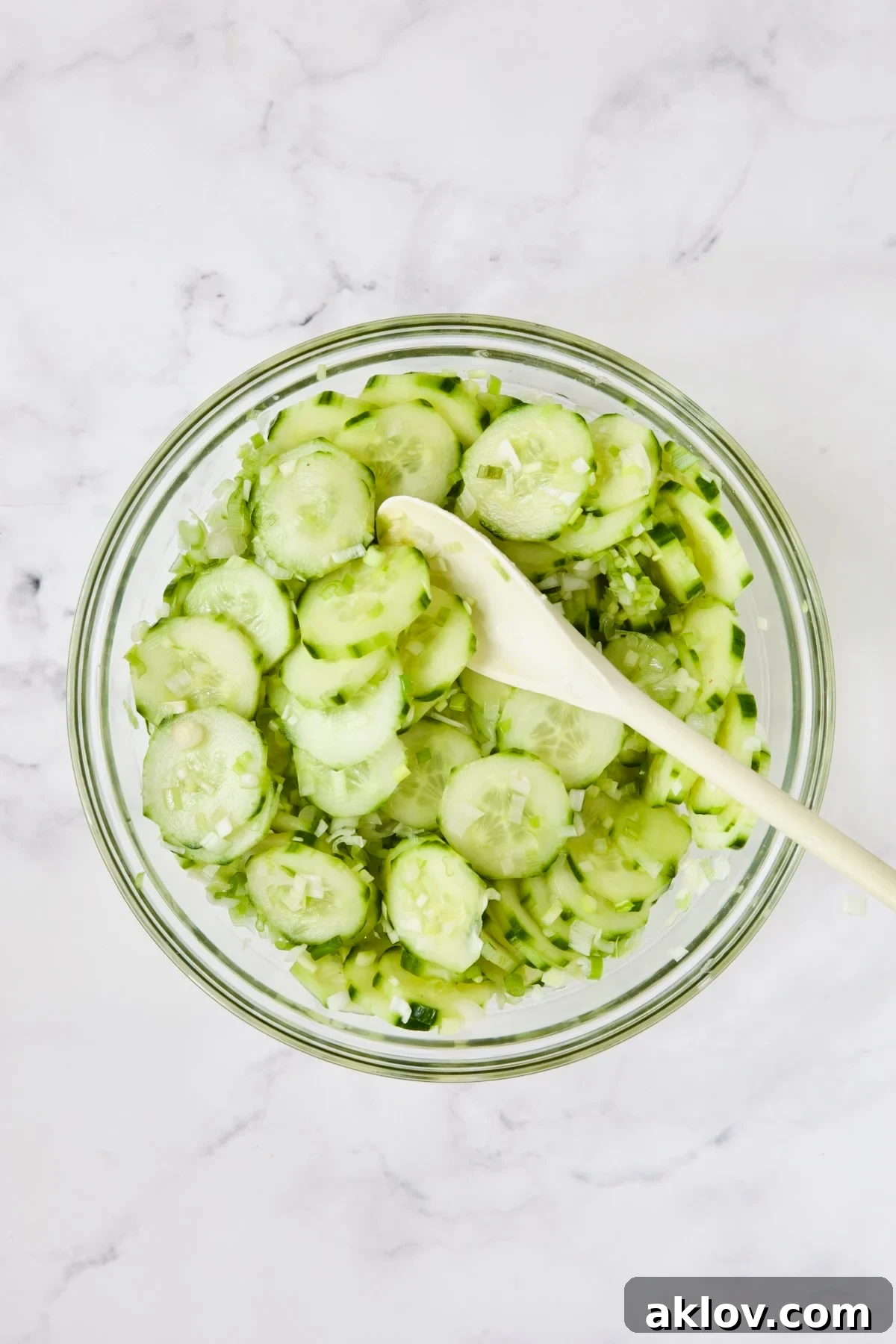 Cucumbers and scallions being stirred with white spoon in glass bowl.