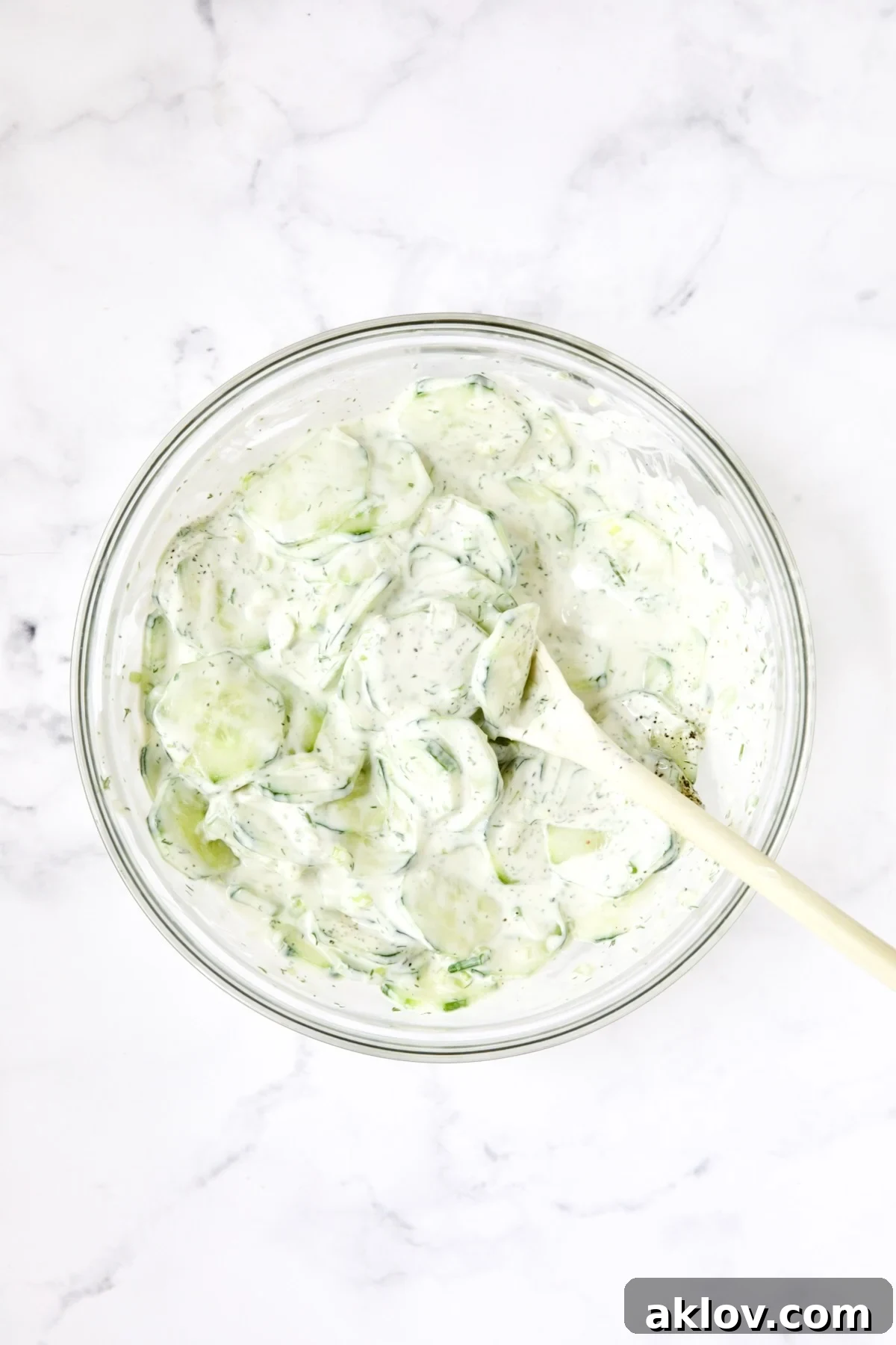Cucumber salad being stirred in glass bowl with white spoon.