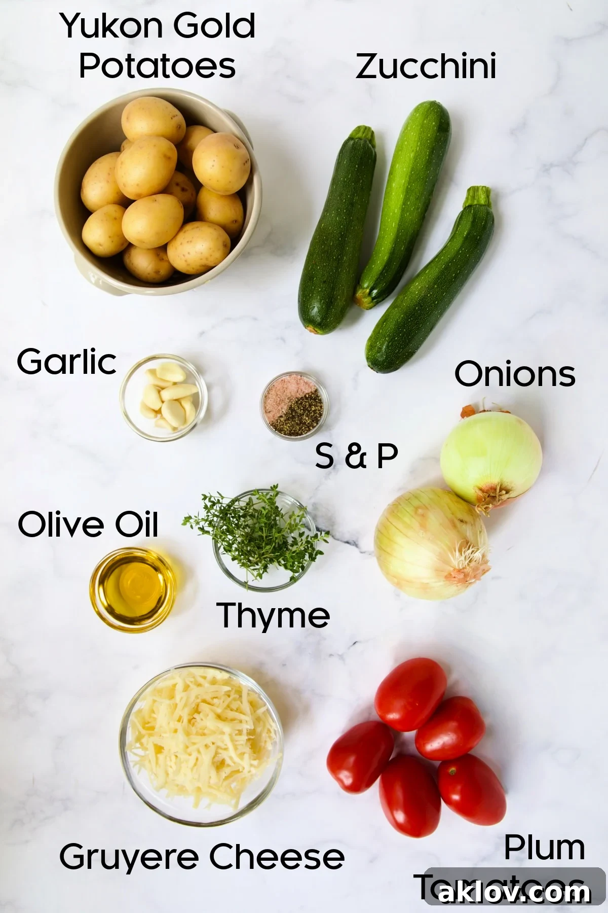 Fresh ingredients for Vegetable Tian laid out on a marble countertop, including sliced potatoes, zucchini, tomatoes, onions, garlic, thyme, and Gruyere cheese.