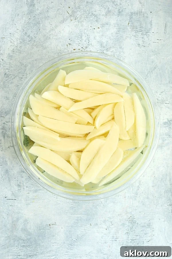Photo of uncooked potato wedges in water in glass bowl.