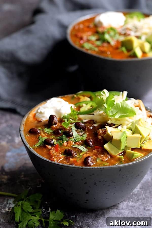 Two bowls of Turkey Black Bean Quinoa Chili, generously garnished with sour cream, shredded cheese, fresh avocado slices, a jalapeno ring, and a sprig of cilantro, ready to be enjoyed.
