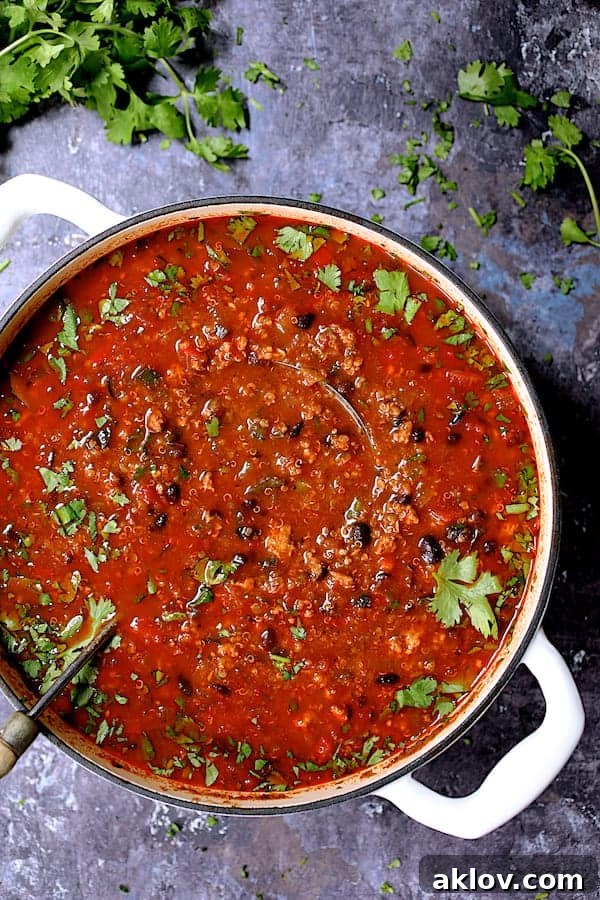 Overhead view of a large white Dutch oven filled with simmering Turkey Black Bean Quinoa Chili, garnished with fresh cilantro, resting on a distressed gray background.