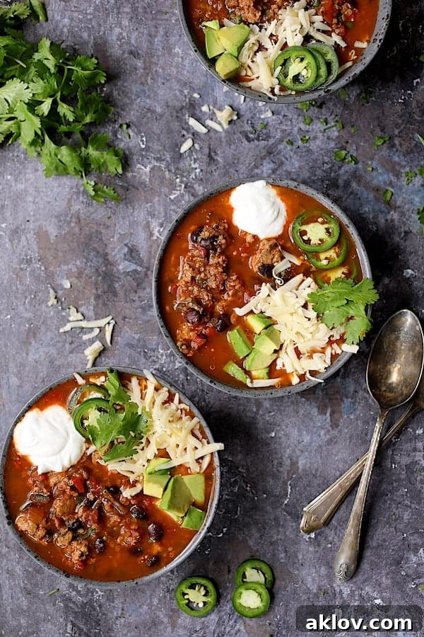 Three rustic gray bowls of Turkey Black Bean Quinoa Chili arranged on a distressed gray background, each beautifully garnished with fresh cilantro, sliced jalapeno, shredded cheese, and diced avocado.