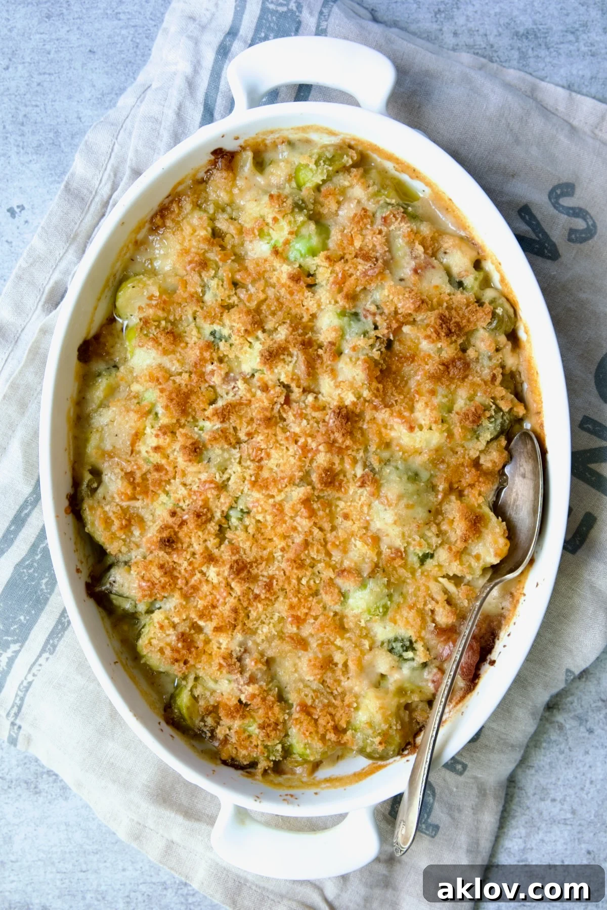 A freshly baked Brussels Sprouts Gratin, bubbly and golden, presented on a blue striped towel with a serving spoon.