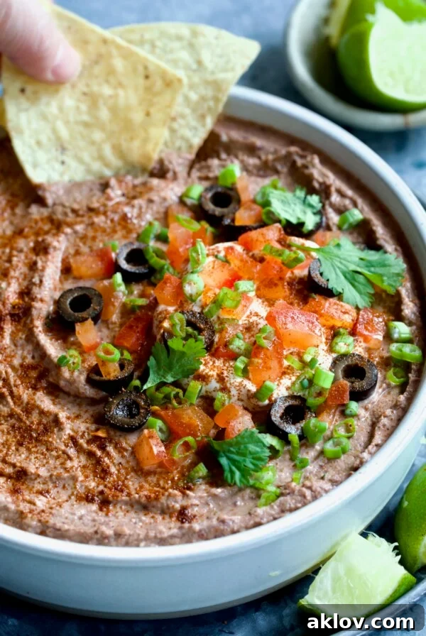 Zesty Black Bean Delight 11 Black Bean Dip garnished with black olives, tomatoes, scallions and cilantro leaves in white clay bowl with tortilla chips and lime wedges.