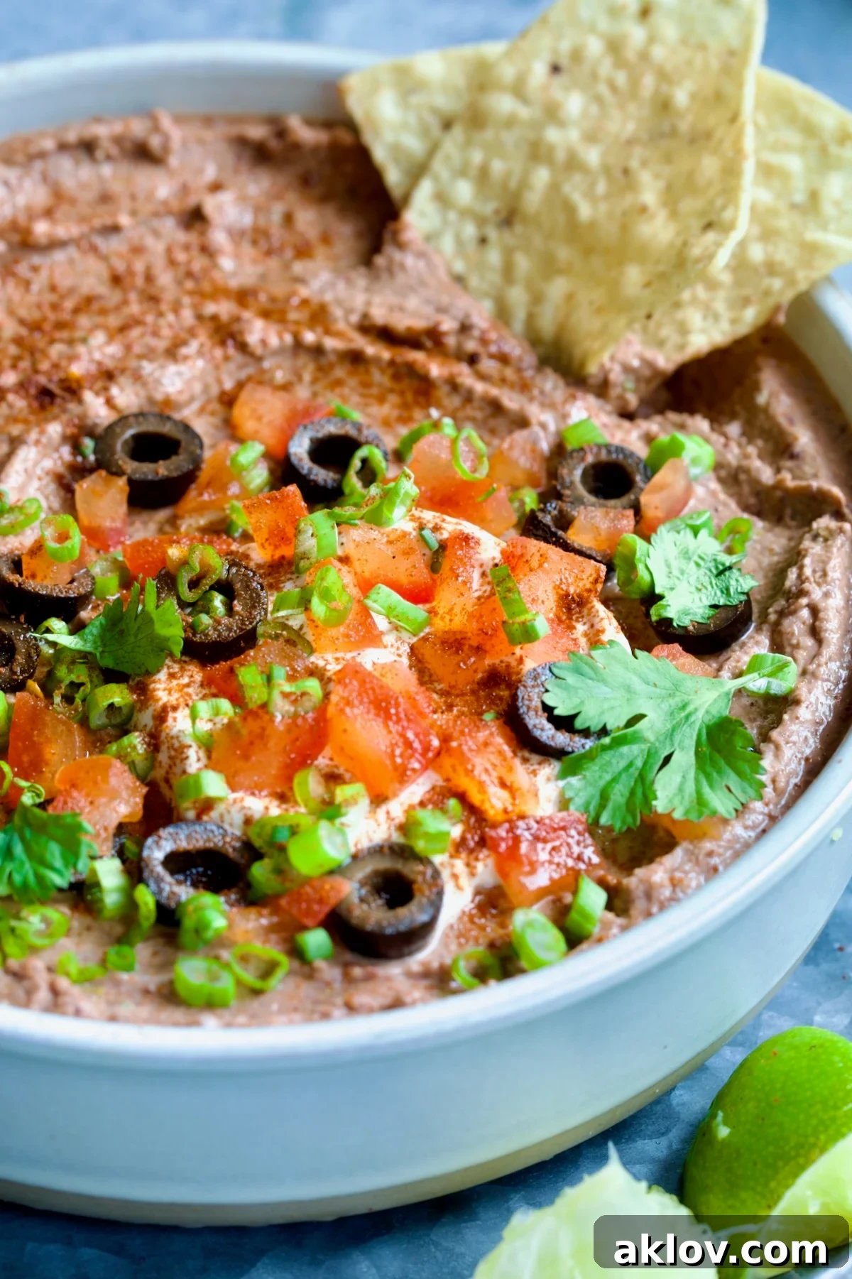 Zesty Black Bean Delight 8 Beautifully garnished Black Bean Dip in a white clay bowl, adorned with black olives, diced tomatoes, green scallions, and fresh cilantro leaves. Tortilla chips and lime wedges are scattered around for an inviting display.