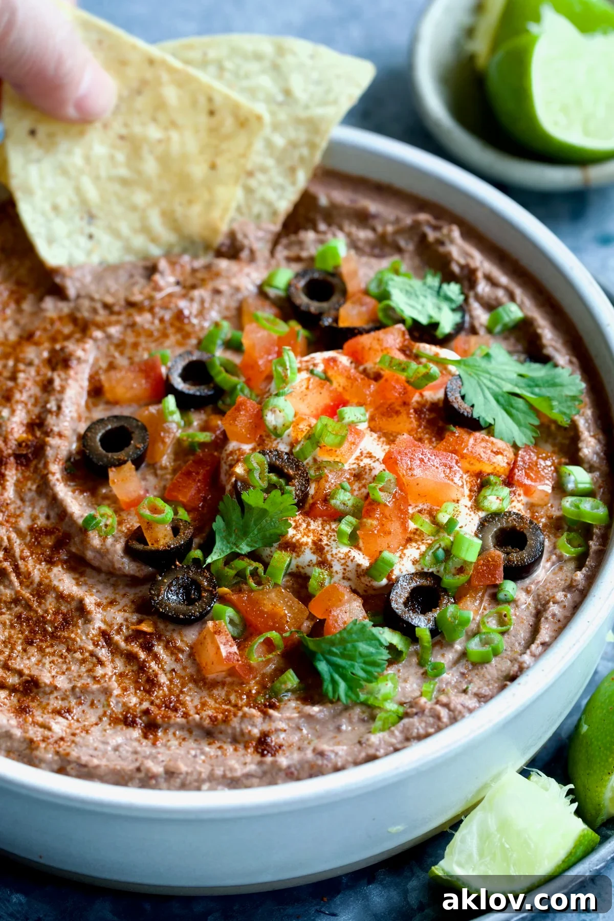 Zesty Black Bean Delight 10 Elegant white clay bowl filled with Black Bean Dip, meticulously garnished with black olives, diced tomatoes, green scallions, and fresh cilantro, complemented by a spread of tortilla chips and lime wedges.