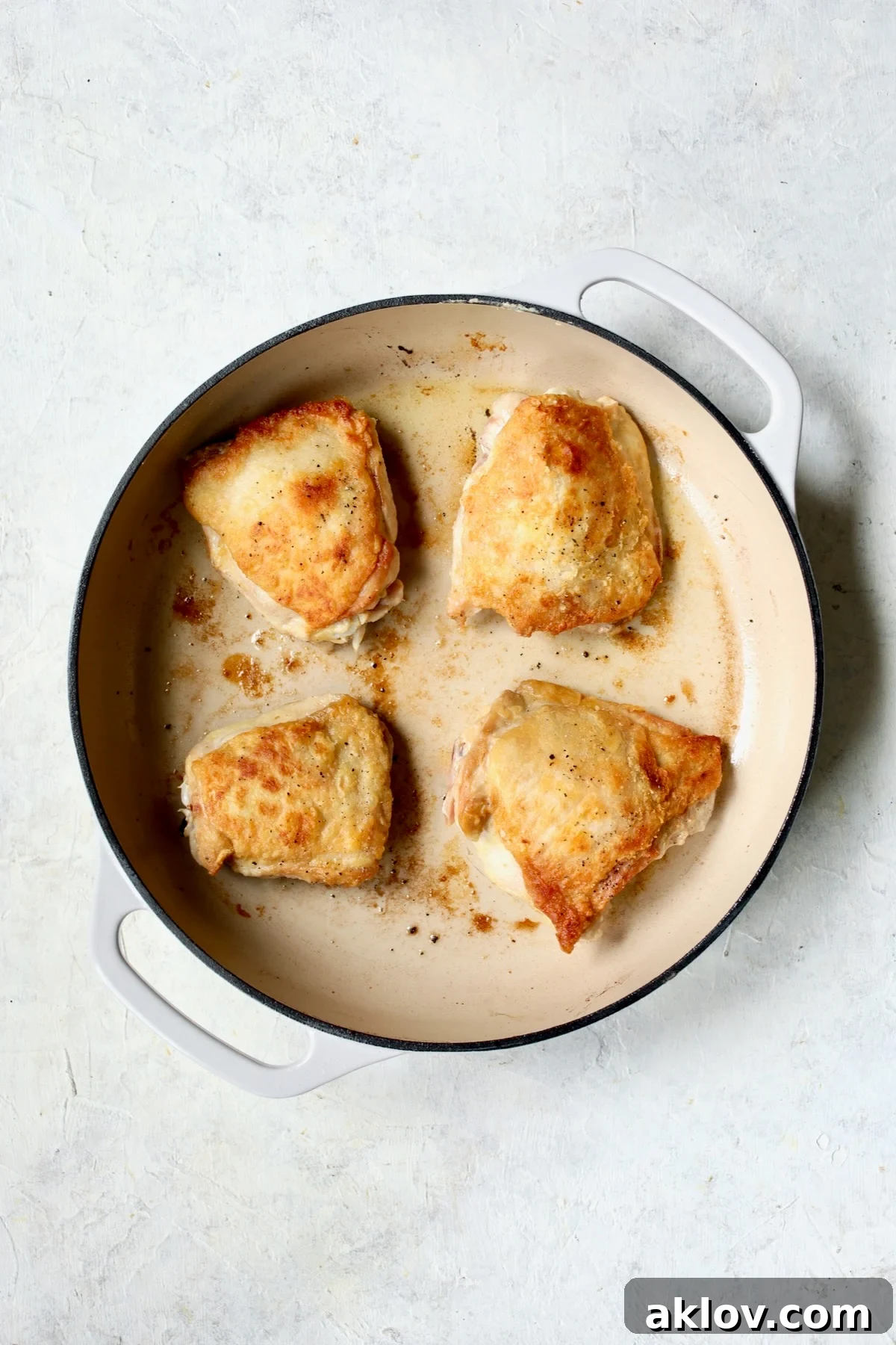 Four browned chicken thighs skin side up in white enamel cast iron skillet.