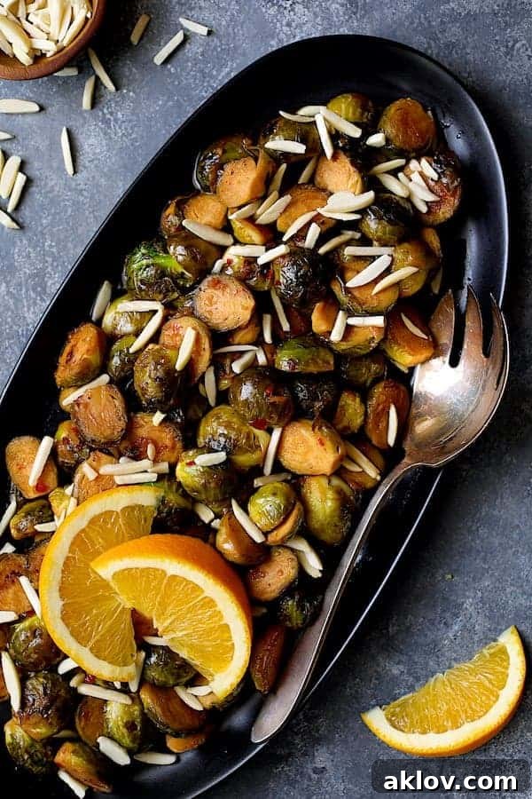 Photo of Pan-Roasted Brussels Sprouts with Thai Sweet Chili Orange Glaze on black platter with serving fork garnished with almonds and orange wedges.