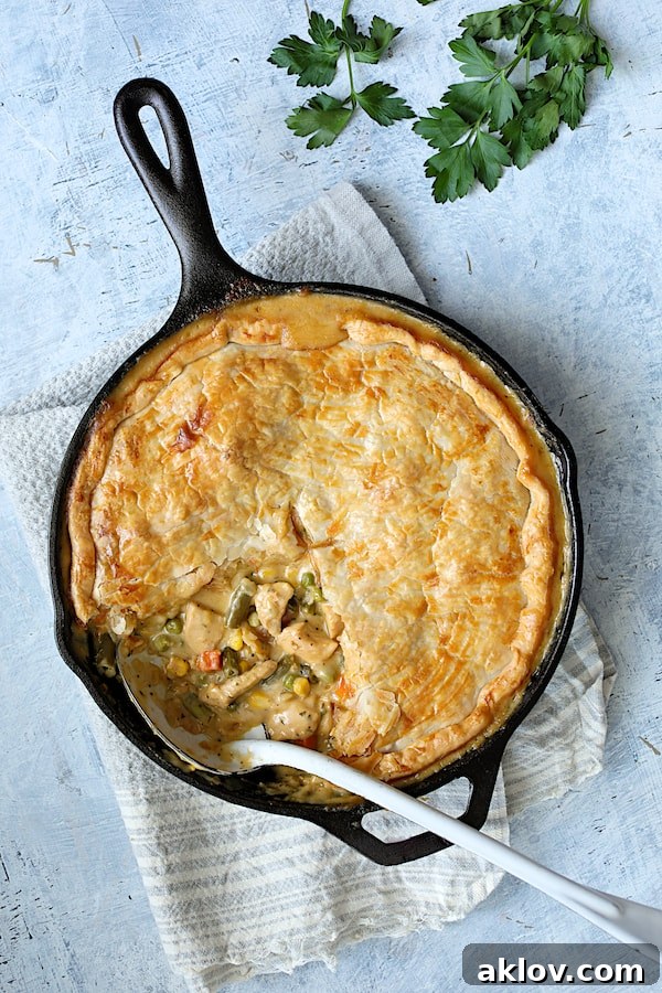 A vibrant photo of Cheesy Skillet Chicken Pot Pie on a light blue background in a cast iron skillet, with a serving removed to expose the rich, creamy filling, tender chicken, and colorful vegetables beneath a golden, flaky crust.