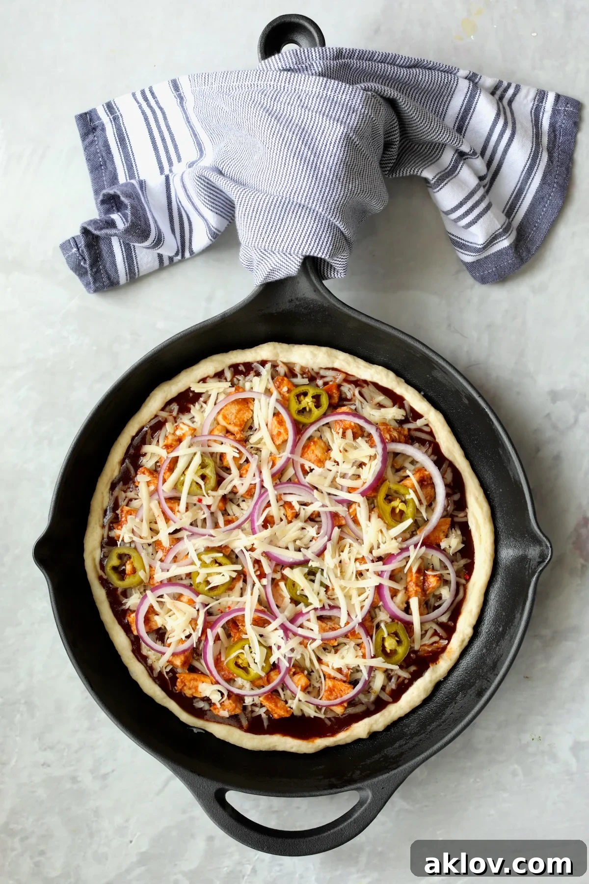 A cast iron skillet pizza fully loaded with barbecue sauce, cheeses, chicken, red onions, and jalapenos, ready for baking.