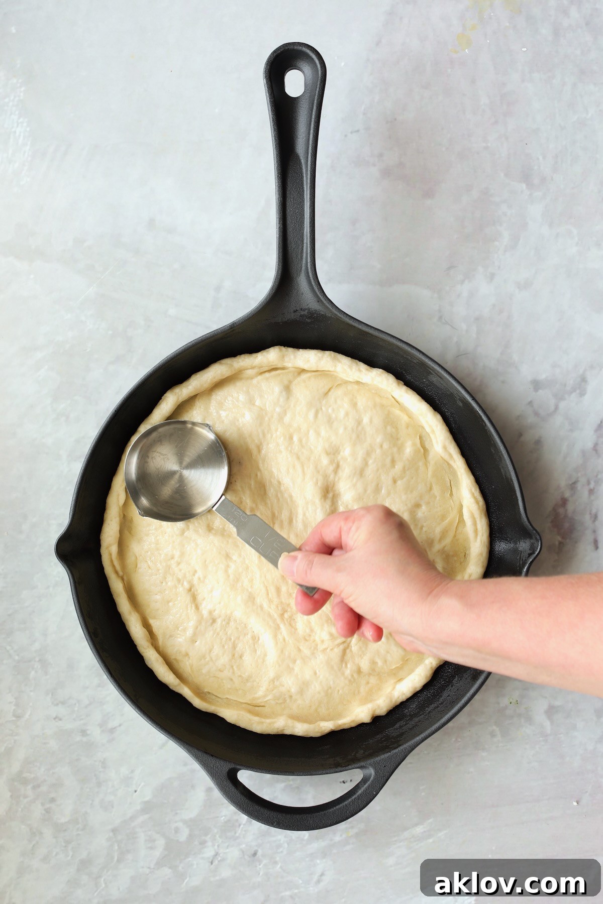 Pizza dough in cast iron skillet being formed to the skillet with 1-cup measuring cup.