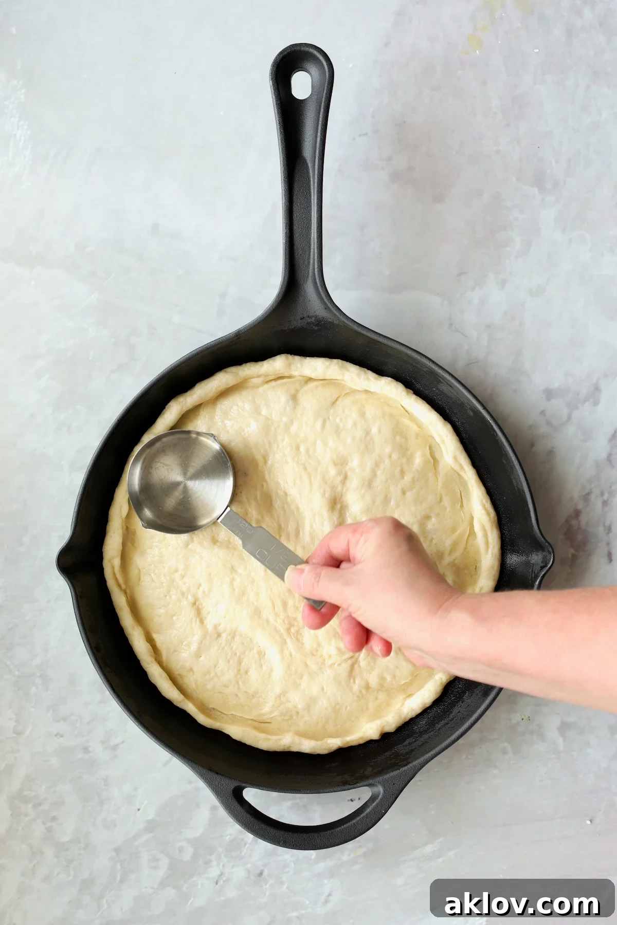 Pizza dough being carefully pressed up the sides of a hot cast iron skillet using a 1-cup measuring cup.