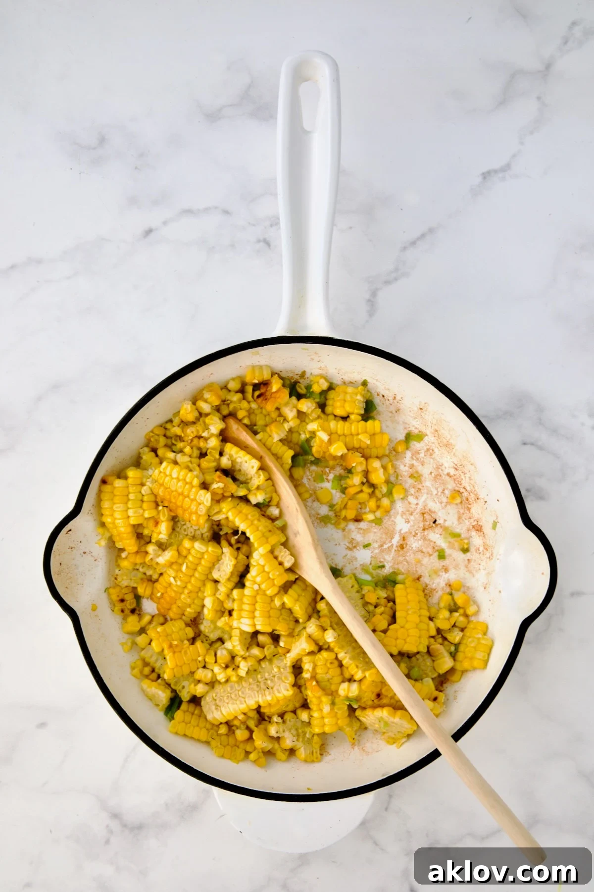 Corn and scallions being stirred with mixing spoon in white enamel cast iron skillet.