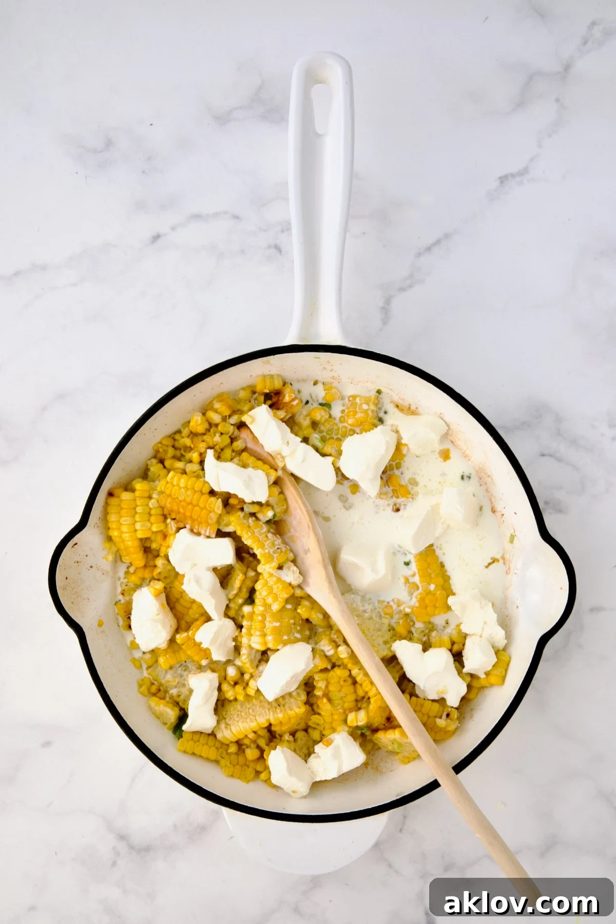 Cream cheese and heavy cream added to corn in white enamel cast iron skillet before being stirred in.