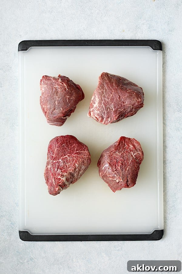 Overhead shot of four beef tenderloin steaks resting on a white cutting board, brought to room temperature.