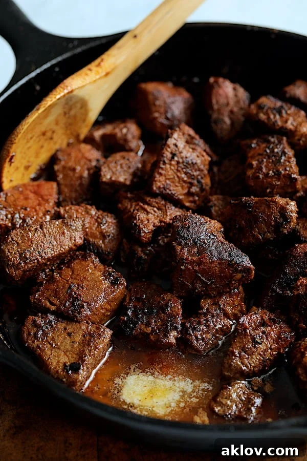Close-up shot of butter being added to the browned steak bites in a cast iron skillet, beginning to melt.