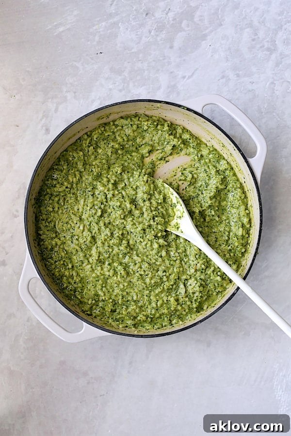 Kale pesto being stirred into the finished risotto in a white cast iron pan with a white spoon, creating a beautiful green swirl.