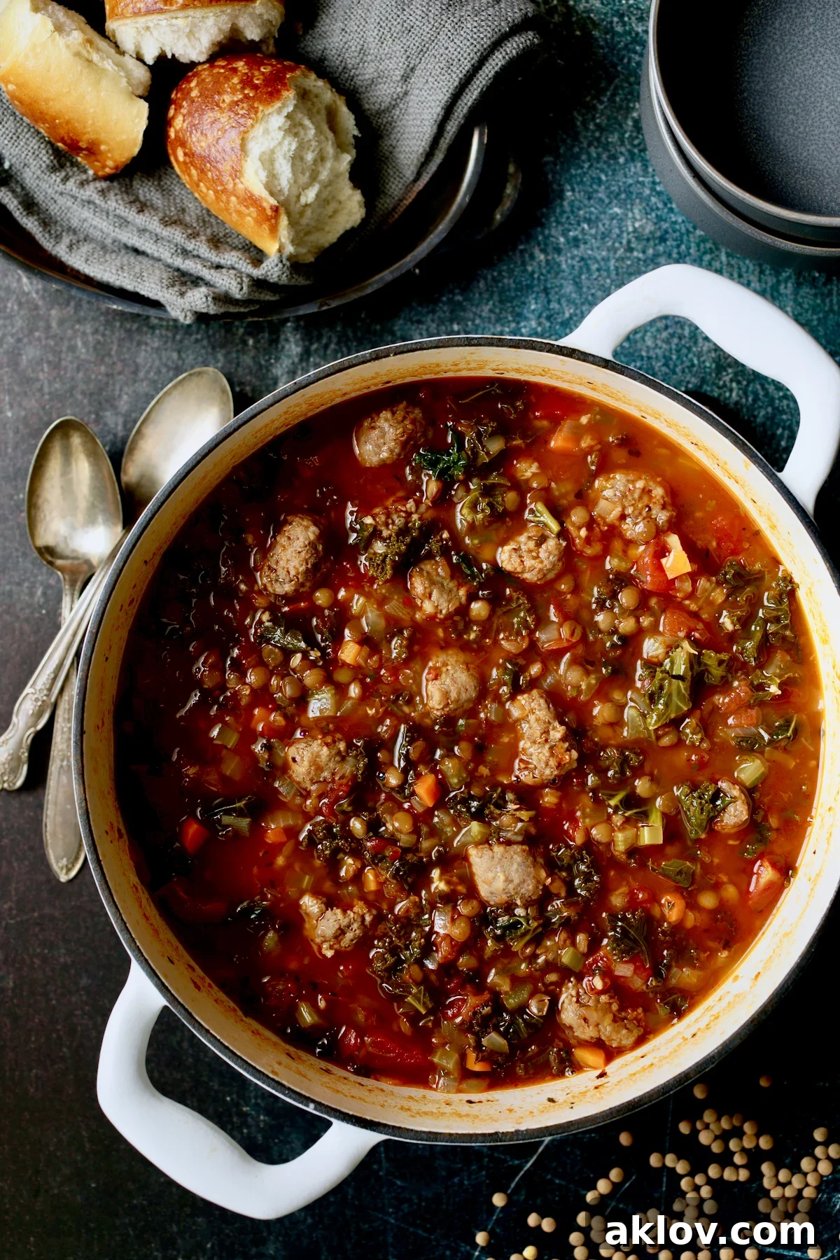 Hearty Sausage and Lentil Soup with Kale served in a white Dutch oven, accompanied by two spoons, two gray bowls, and fresh bread on a gray napkin, embodying comfort food.