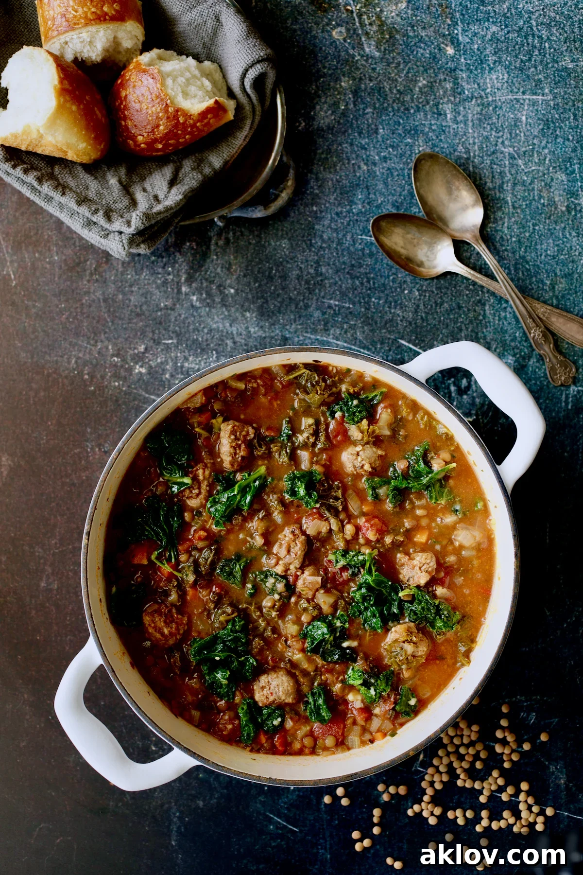 Sausage and Lentil Soup with Kale in a white Dutch oven, served with fresh bread, two spoons, and scattered lentils, highlighting its inviting presentation.