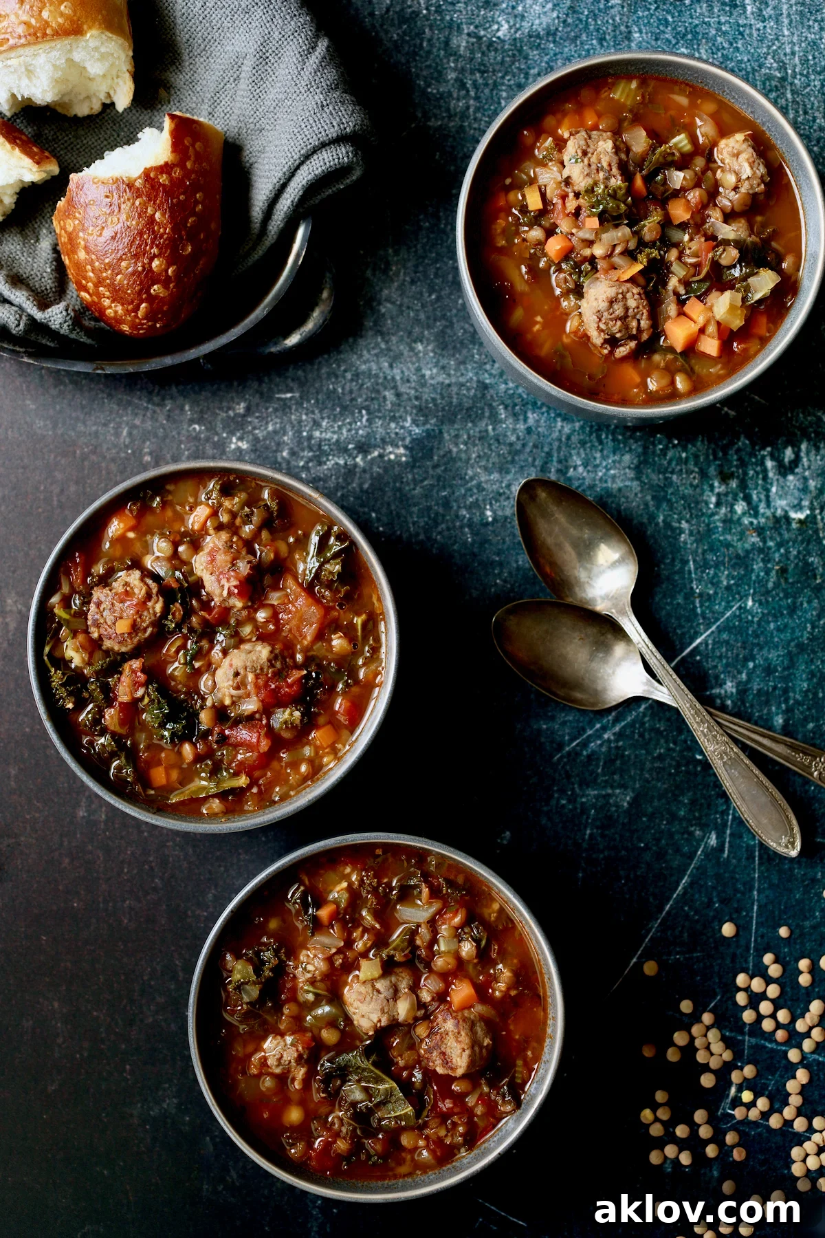 Three inviting bowls of Sausage and Lentil Soup with Kale, accompanied by bread on a gray napkin, ready for a comforting meal.