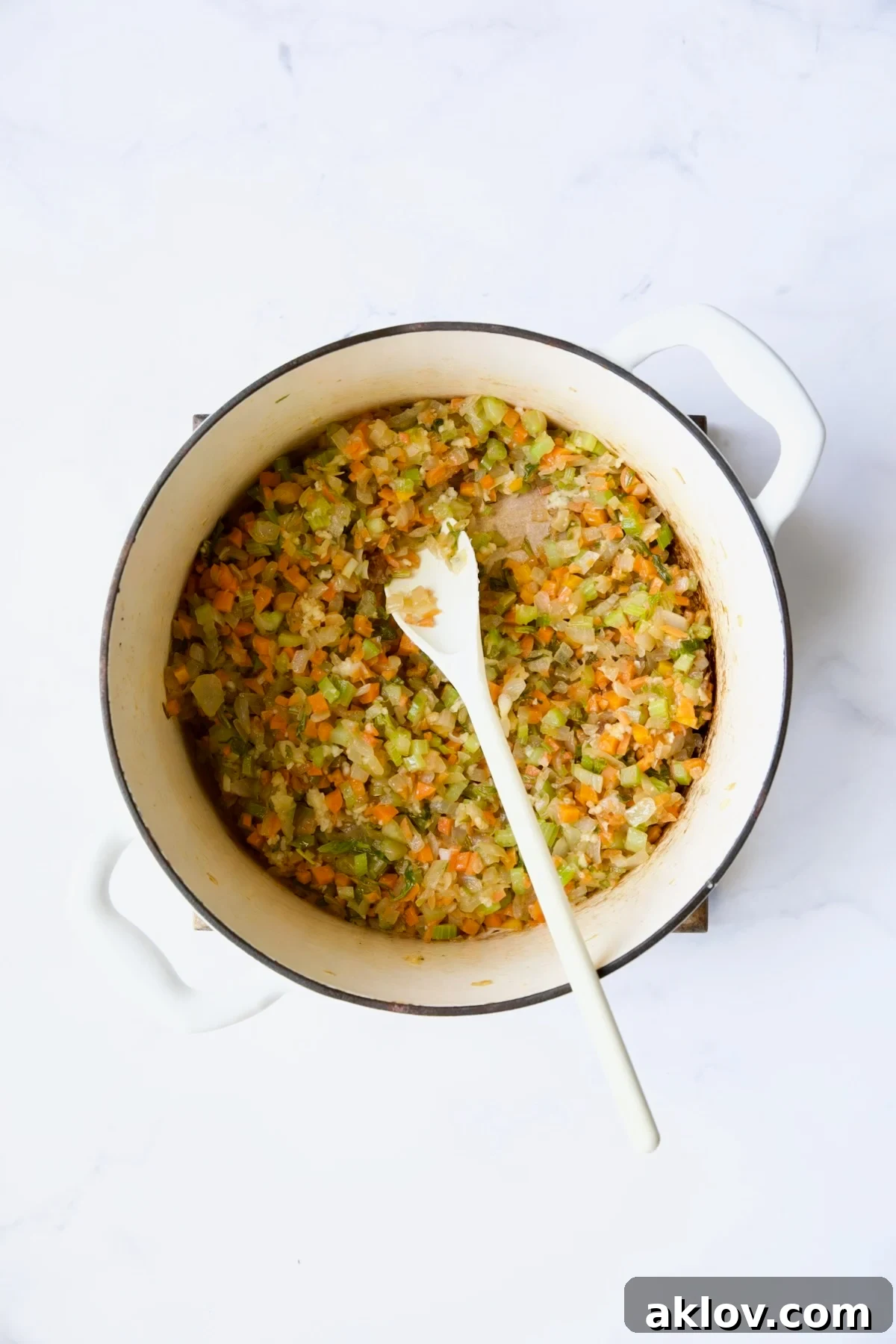 Sautéing chopped onions, celery, and carrots in a white Dutch oven, stirred with a white spoon.