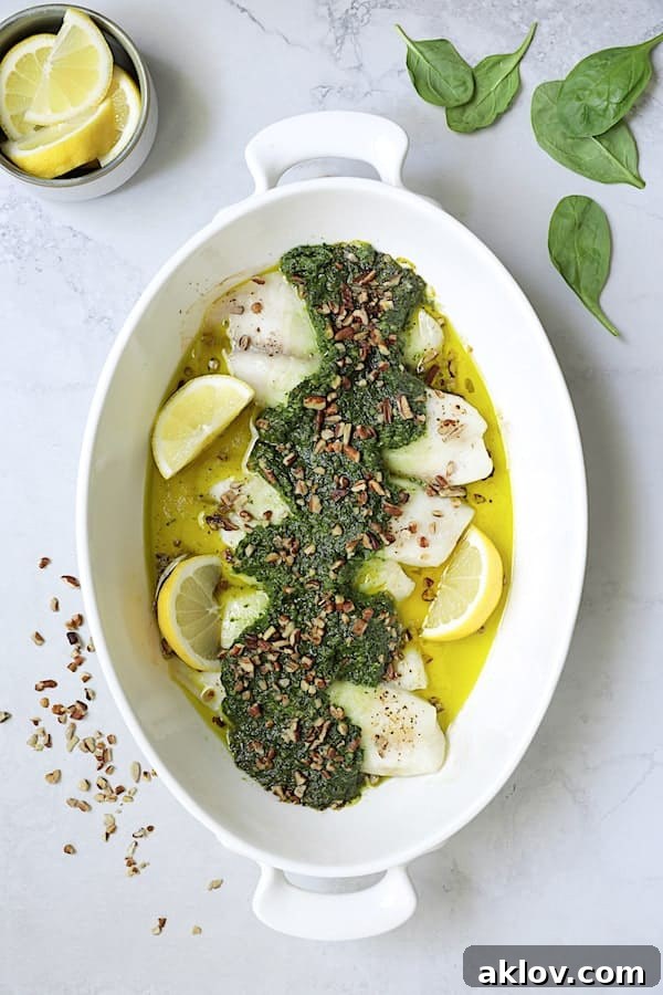 Photo of Baked Tilapia with Spinach Pecan Pesto in white oval baking dish garnished with lemon wedges and chopped pecans. A complete and healthy meal ready to serve.