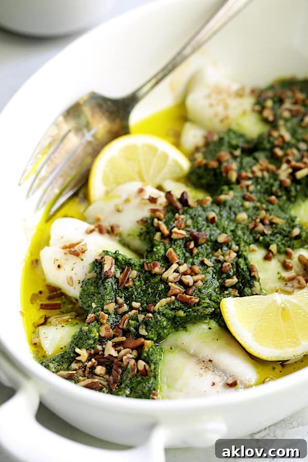 Close-up photo of Baked Tilapia with Spinach Pecan Pesto in white baking dish garnished with chopped pecans and lemon wedges. The pesto topping looks rich and flavorful.
