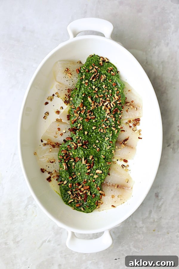 Photo of uncooked tilapia fillets in a baking dish, generously topped with green pesto and sprinkled with chopped pecans.