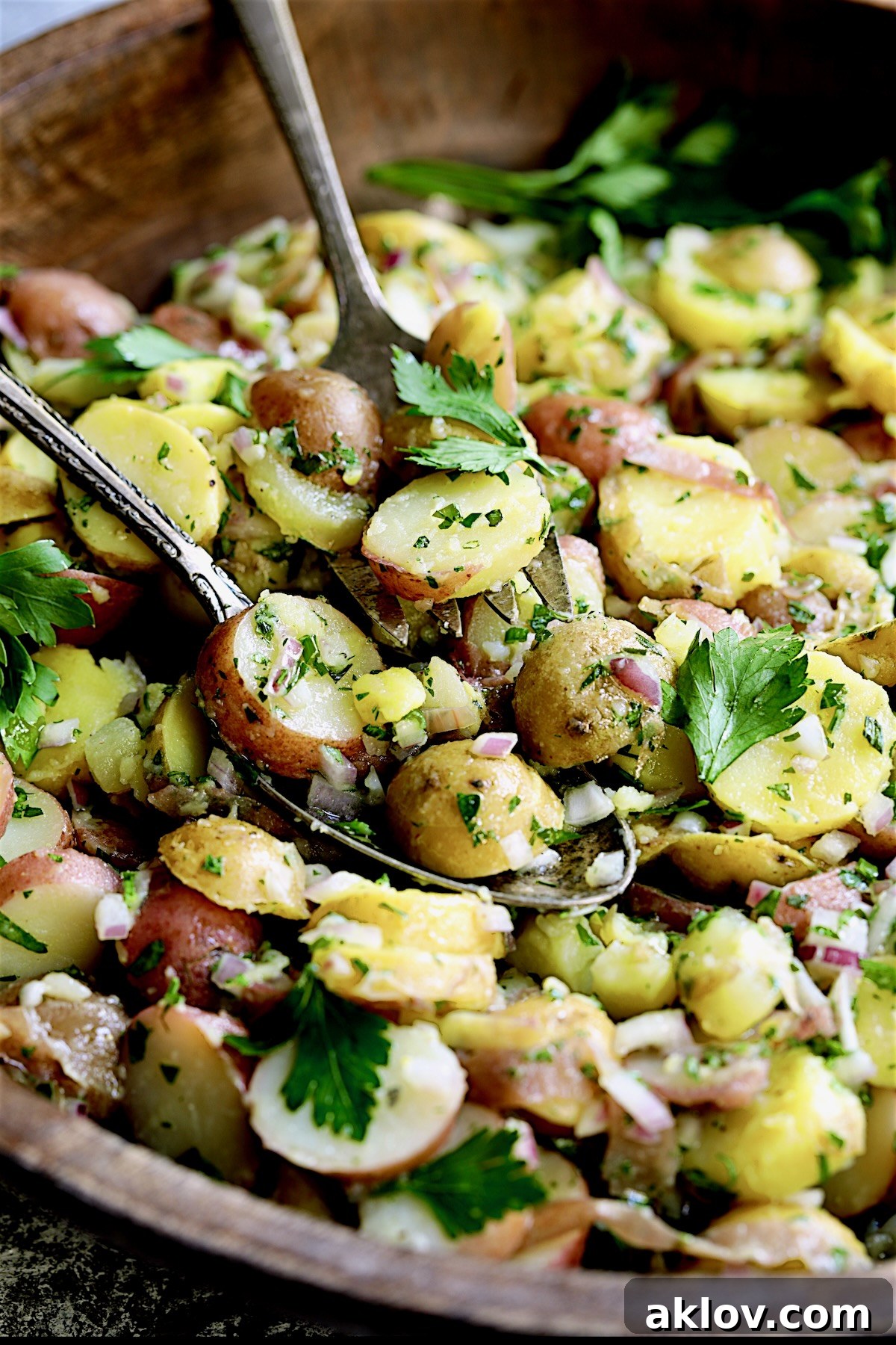A close-up of French Potato Salad being served from a wooden bowl with elegant serving utensils, highlighting its fresh ingredients.