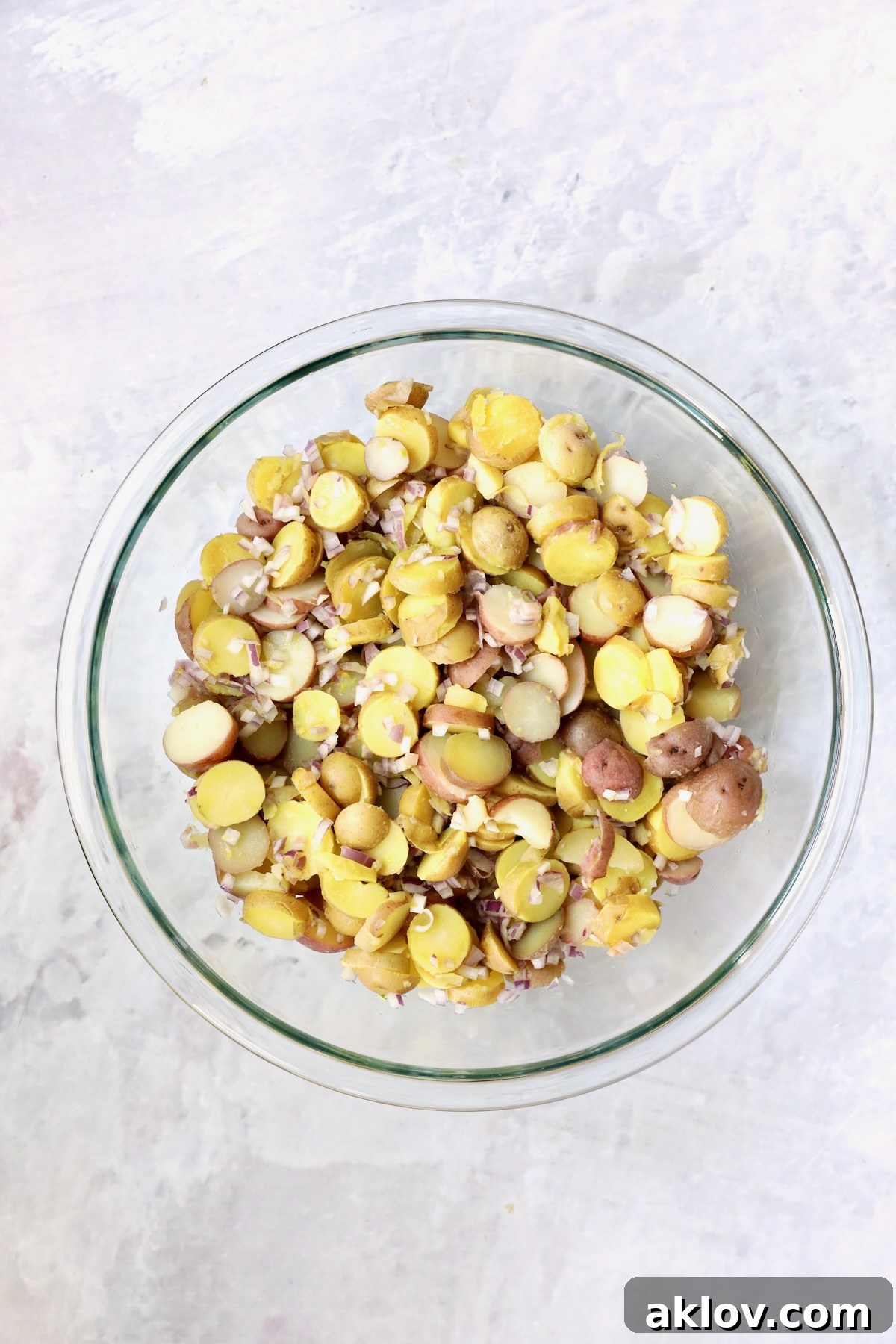 Sliced, cooked red and yellow new potatoes mixed with minced shallots in a glass bowl, absorbing the wine and broth mixture.