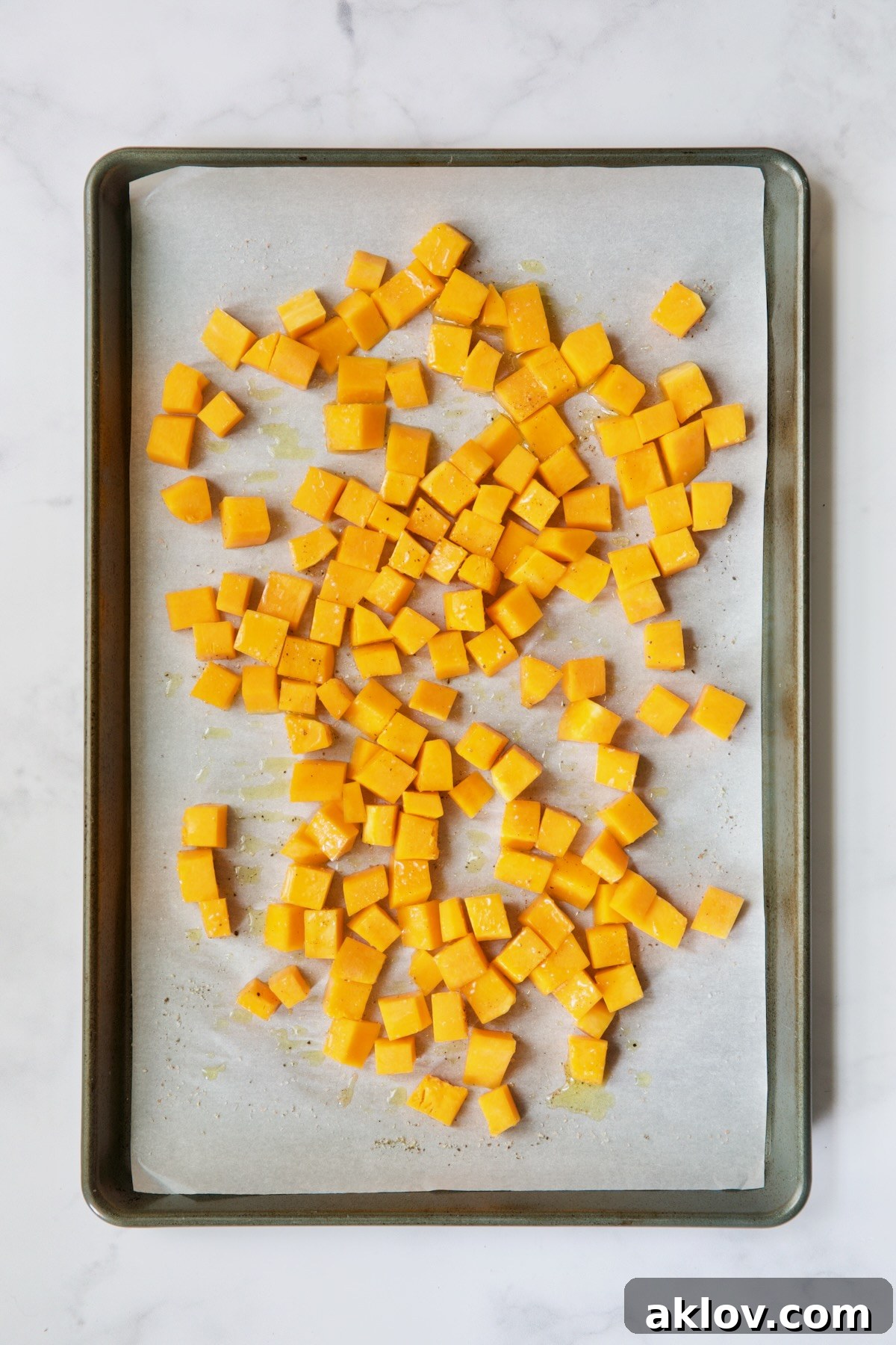 Butternut squash cubes, tossed with olive oil and seasoning, spread on a white parchment paper-lined baking sheet, ready for roasting.