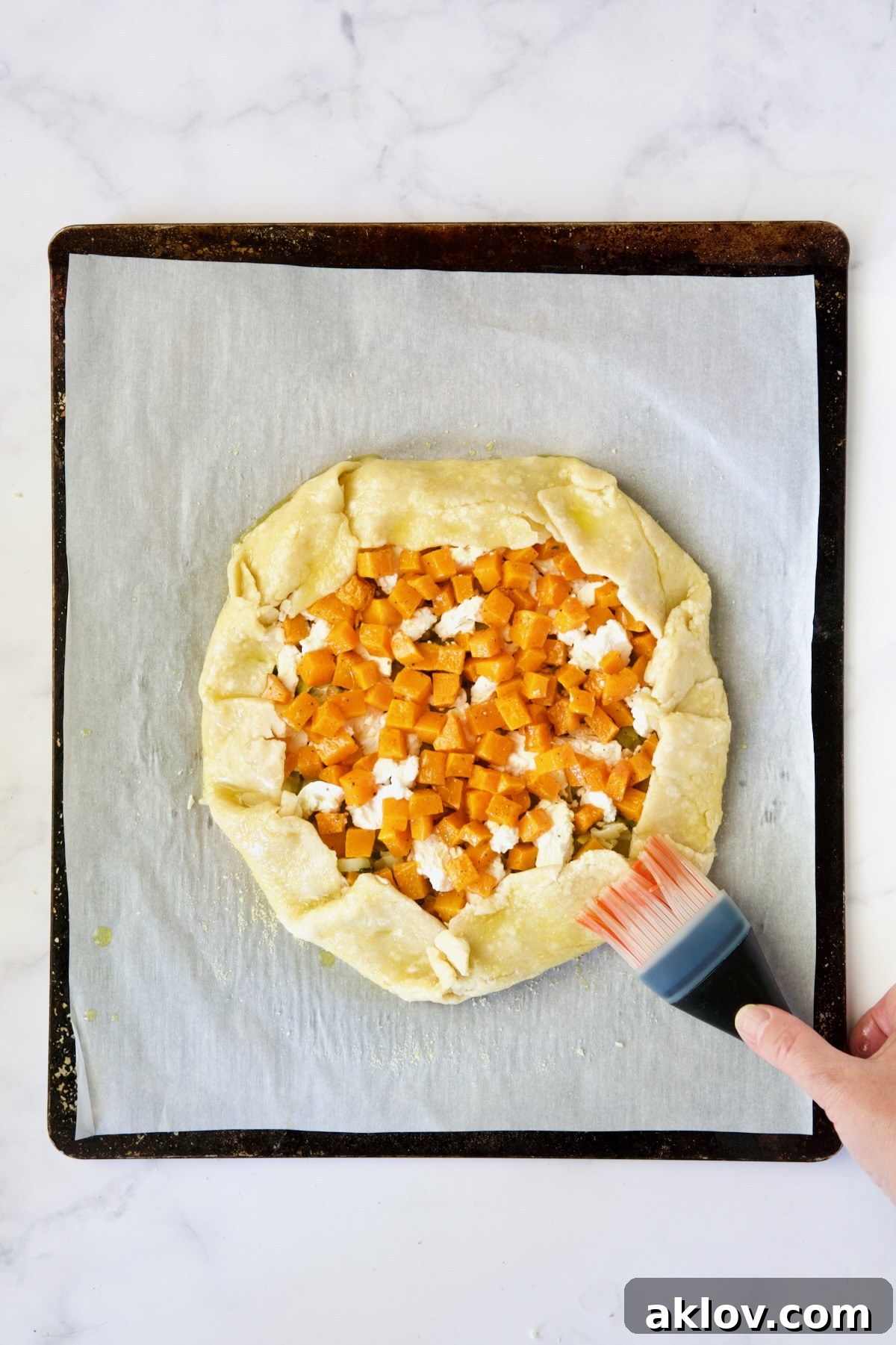 The assembled galette on a white parchment paper-lined baking sheet, with its rustic edges folded over the filling and being brushed with olive oil for a golden crust.