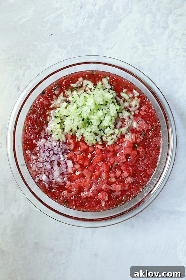 Remaining diced ingredients, including small watermelon cubes, cucumber, red onion, and serrano chiles, being added to the pureed watermelon base in a large glass bowl for the gazpacho.