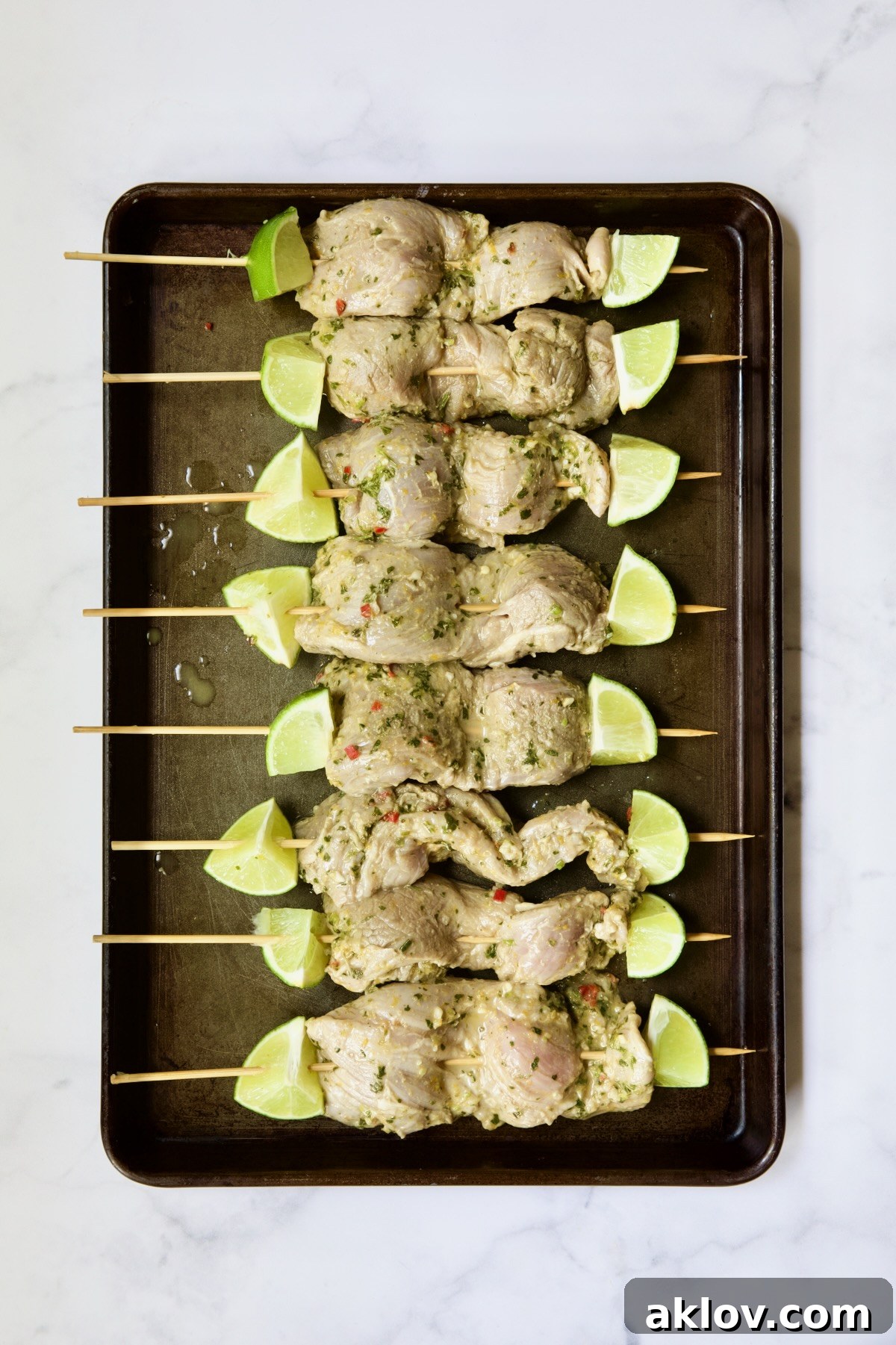 Uncooked Mojo-Marinated Pork Tenderloin Skewers with lime wedges, arranged on a rimmed baking sheet.