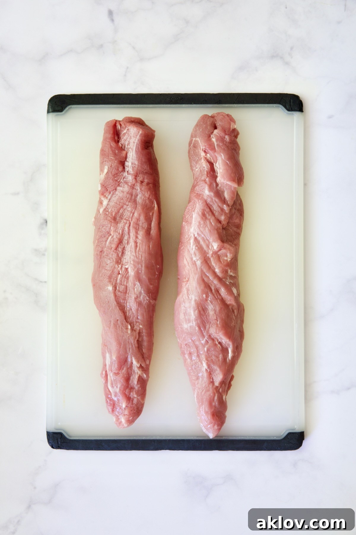 Two trimmed pork tenderloins on a white plastic cutting board, ready for slicing.