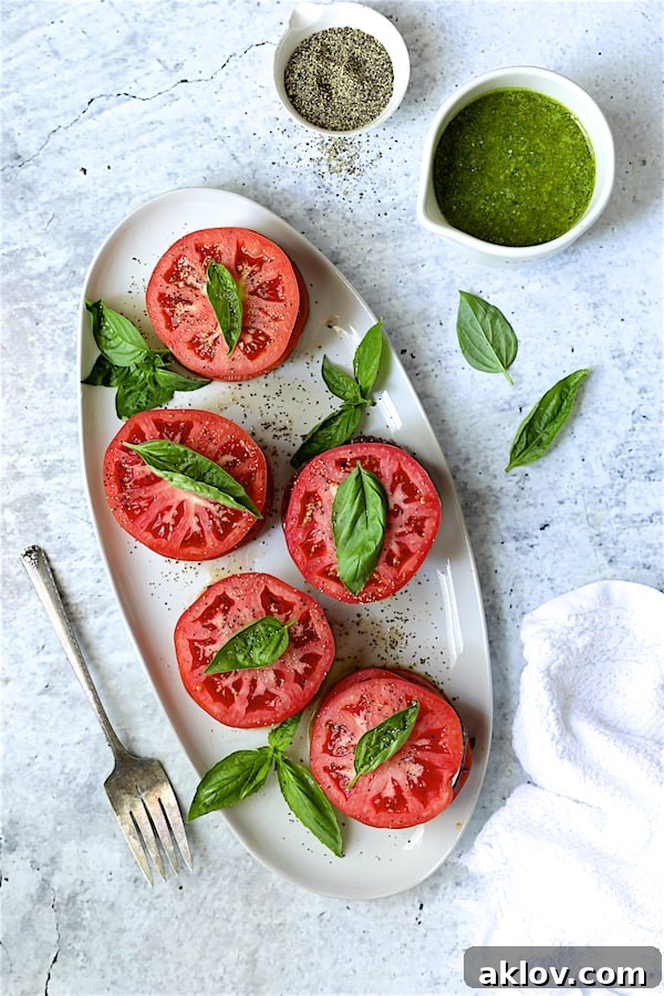 Five beautifully assembled stacked tomato salads, garnished with fresh basil, arranged on a white oval platter, ready to be drizzled with dressing and served.