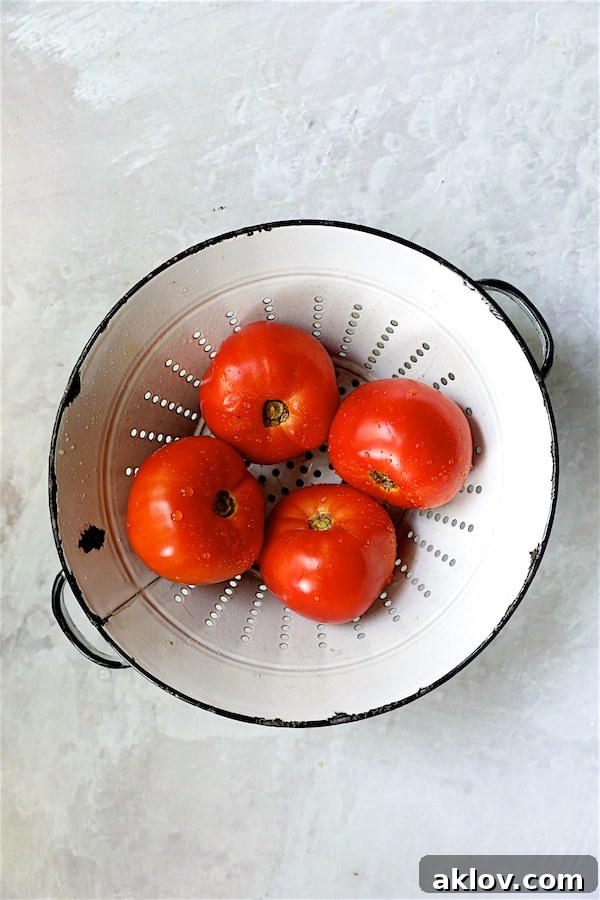 Four perfectly ripe, red tomatoes displayed in a rustic white colander, ready to be sliced for the salad.