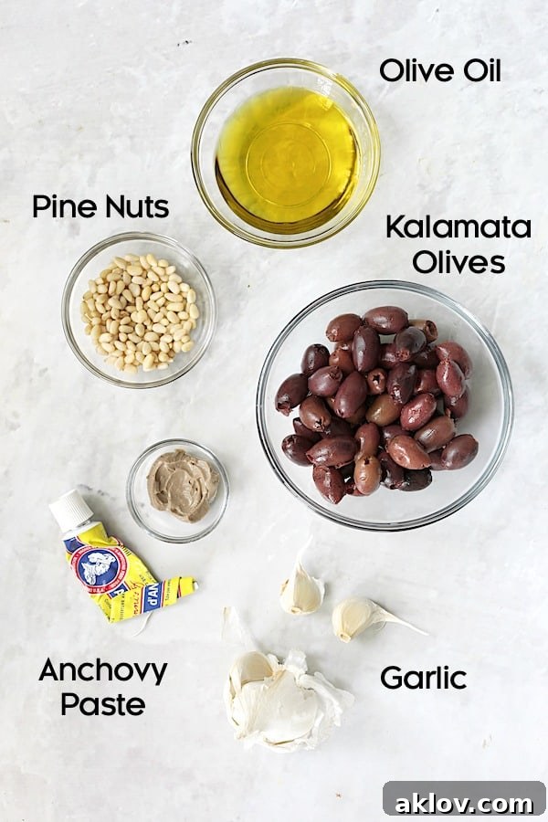 Individual glass bowls containing the fresh ingredients for black olive tapenade: pitted Kalamata olives, garlic cloves, pine nuts, and a tube of anchovy paste.