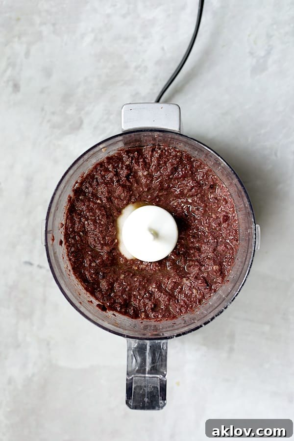 The finished black olive tapenade, a rich, dark spread, visible inside the bowl of a food processor after blending.