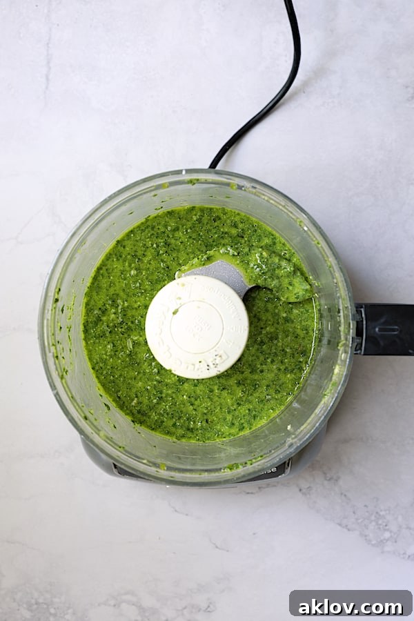 The freshly blended basil pesto dressing, a vibrant green, swirling inside the bowl of a food processor.