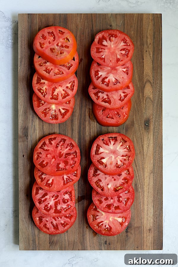 Sixteen freshly cut, even slices of ripe red tomatoes arranged neatly on a wooden cutting board, ready for assembly.