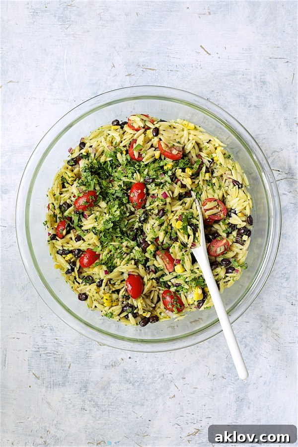 A white spoon gently stirring the Southwestern Orzo Salad ingredients within a clear glass bowl, showcasing the dressing being incorporated.