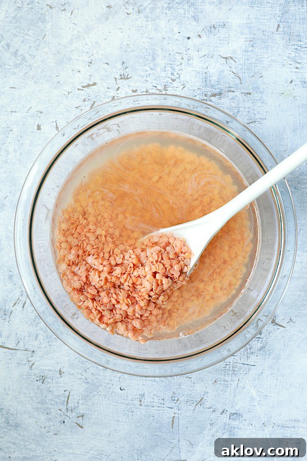 Red lentils soaking in a clear glass bowl, being gently stirred with a white spoon to ensure even hydration.