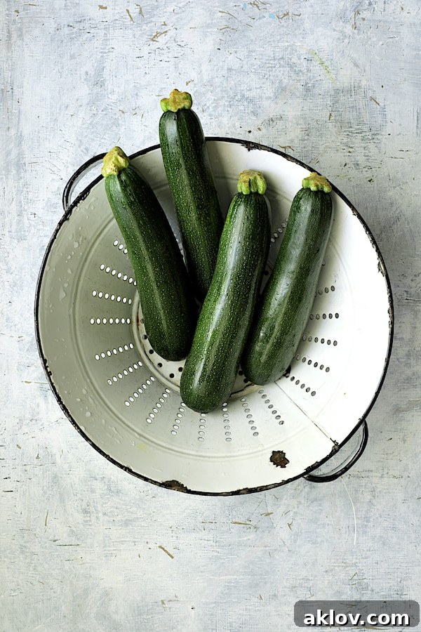 Four fresh, green zucchini nestled in a white distressed colander, showcasing their readiness for shredding.