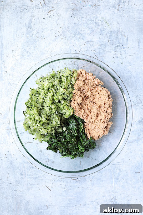 A colorful mixture of squeezed zucchini, pureed red lentils, and sautéed spinach in a large glass bowl, ready to be formed into fritters.