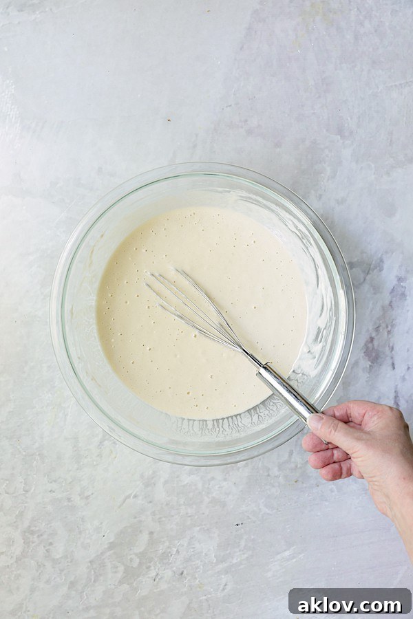 A wire whisk stirring the beer batter in a clear glass bowl, showing its smooth consistency.